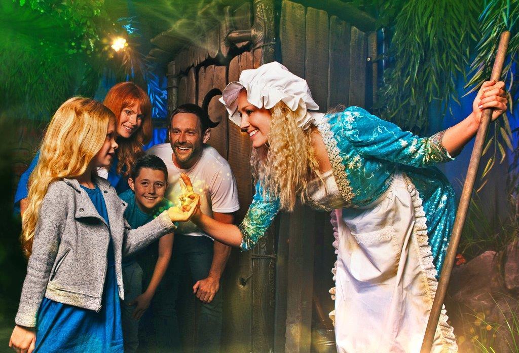 A female actor is dressed as a maid at Shrek's World of Adventure. She is smiling at a guest who is a young blonde child. There is a green sparkle of magic between their hands.