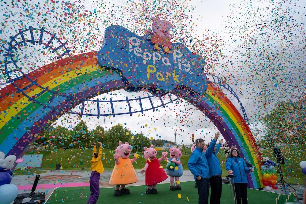 A rainbow sign with a blue title 'Peppa Pig Park' with a pink animated pig above it. Underneath the sign there is a group of actors dressed up as the Peppa Pig family. In the sky there is colourful confetti.