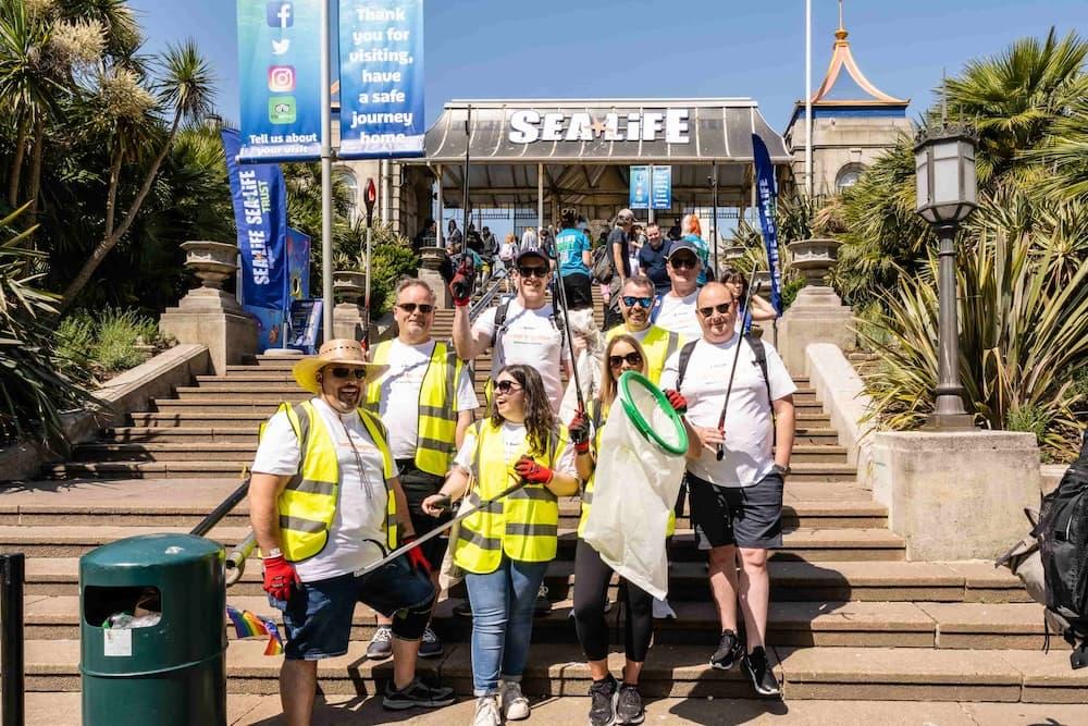 A group of SEA LIFE Trust employees are wearing bright yellow high-vis jackets. They are holding litter picks and bin bags outside of a beach.