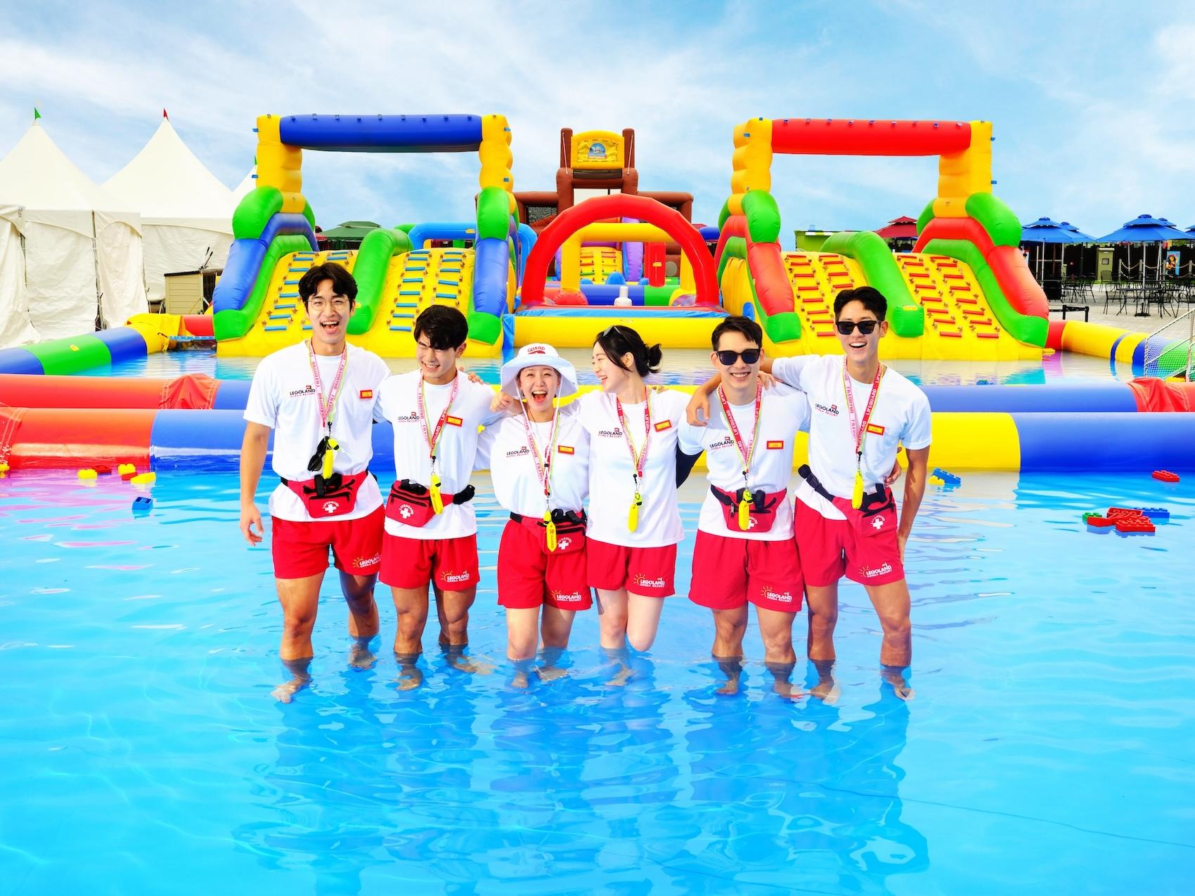 Six LEGOLAND lifeguards stand in knee-depth pool water. Behind them is a gigantic inflatable obstacle course.