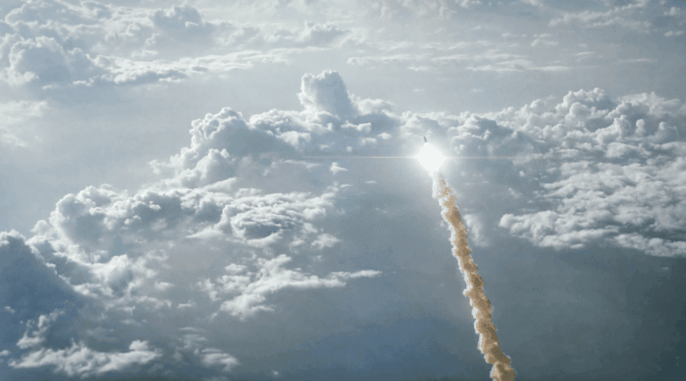 A rocket is going through the clouds leaving a trial of smoke behind it. The sun is shining and the background is filled with white clouds.