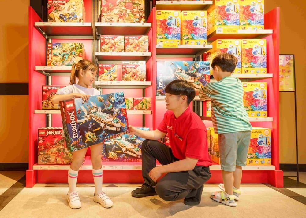 LEGOLAND® employee kneels next to two children in front of LEGO® shelves. The employee helps a girl with a LEGO box.