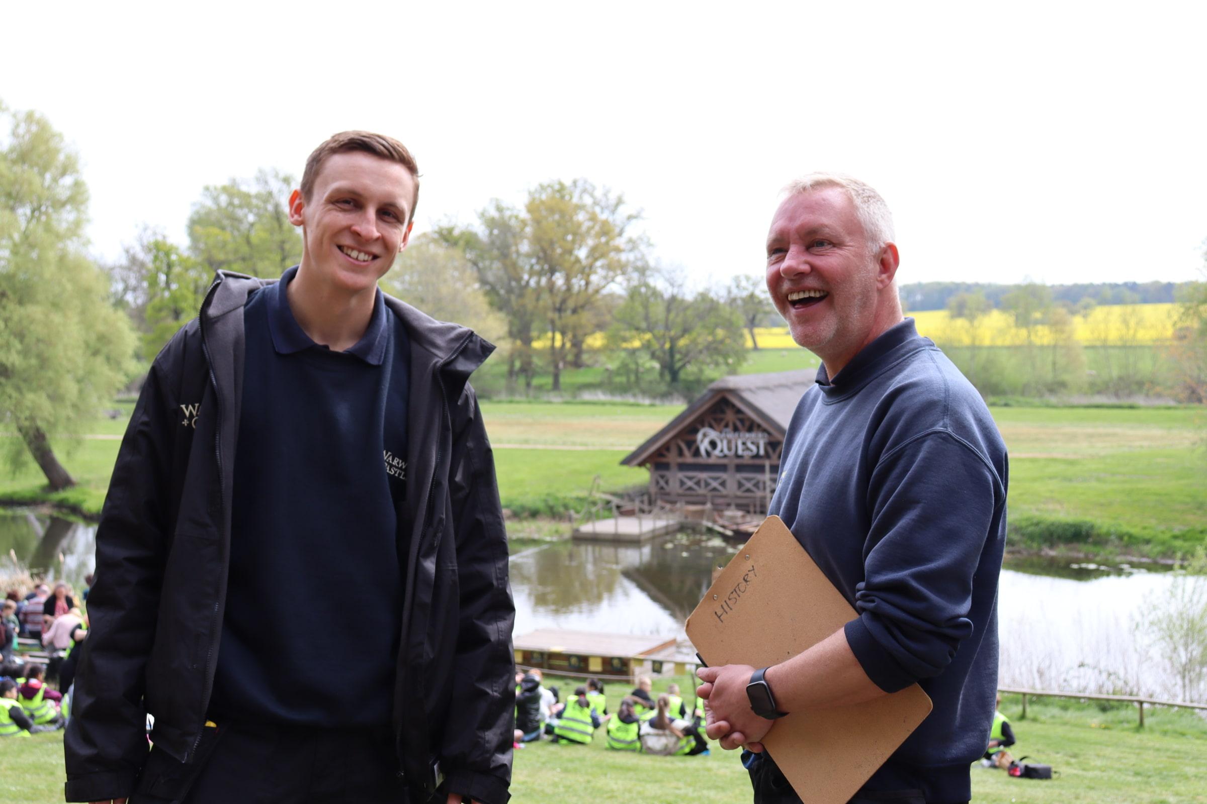 Two men stand outside infront of water, one smiles at the camera with the other laughing on an angle holding a clipboard.