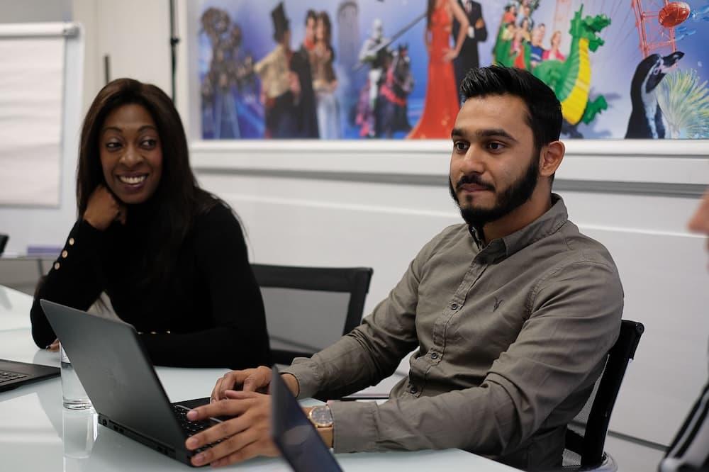In a white office a woman sat next to a man smiles and looks towards him as he is on his laptop looking past the camera.