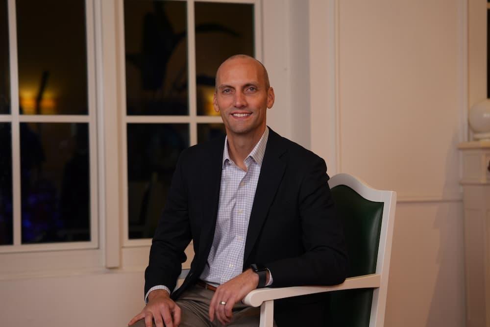 A man in a black blazer and white shirt smiles on a leather chair in front of a white wall. A window behind him is blacked out.