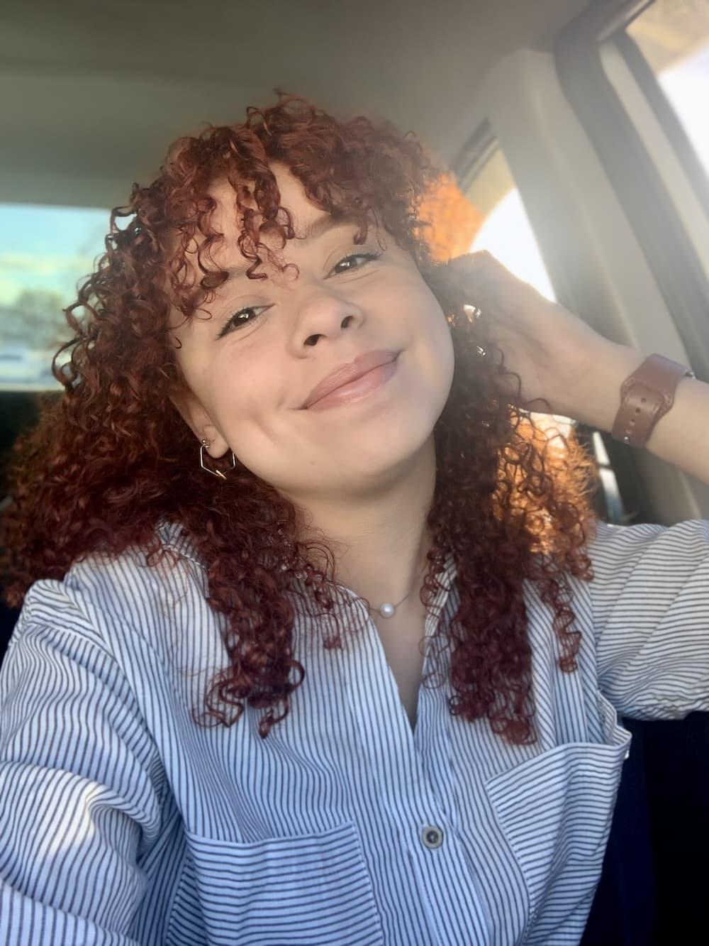 Women with curly, auburn hair is wearing a blue and white striped shirt. She is smiling at the camera for a selfie.