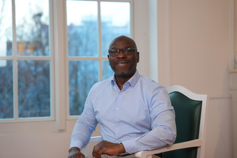 Man in a blue shirt sits on a green leather chair in front of a white wall. Blurred in the background is a window.