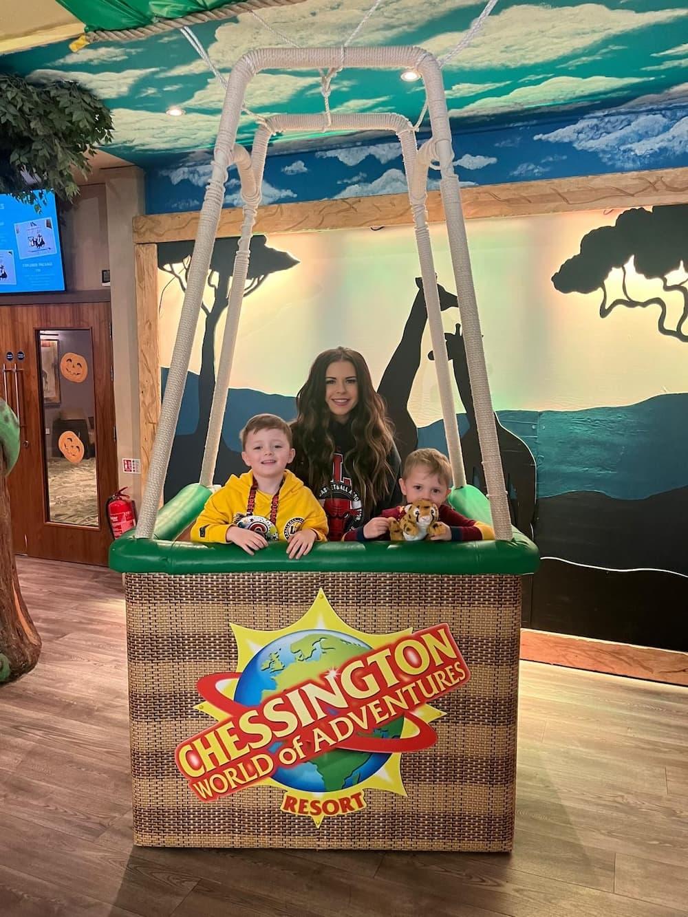 Hot air balloon basket indoor with the Chessington World of Adventures Resort logo holds two children and a woman. There is a landscape adventure backdrop.