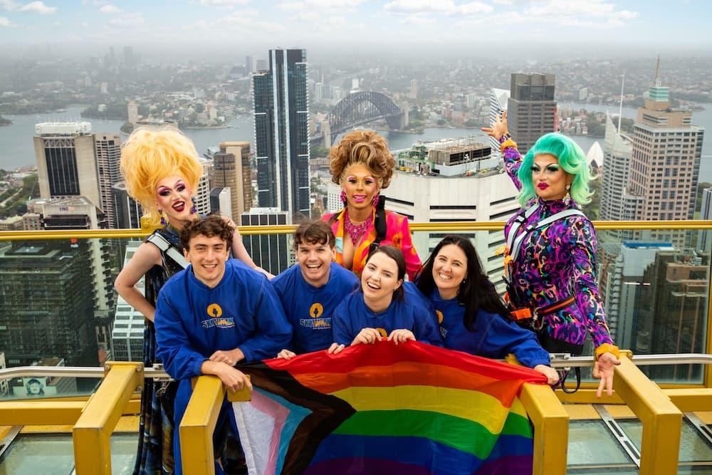 Four employees are wearing blue Sydney Tower Eye uniform and are crouched down at the top of the tower holding a pride flag, with three drag queens behind them. In the background there are buildings and the skyline of Sydney.