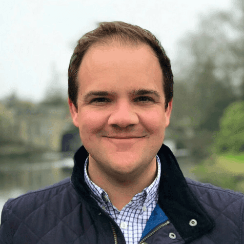 Man with short brown hair is wearing a checked blue and white polo shirt with a blue quilted coat. In the background, there is a river and country landscape.