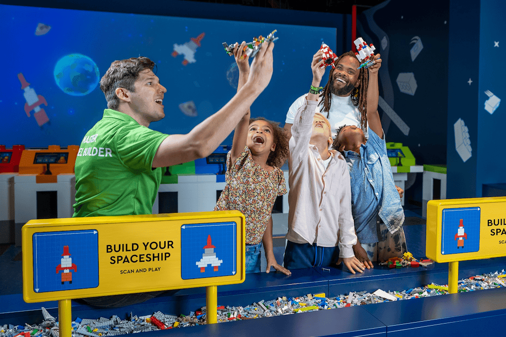 A model builder is wearing a green polo shirt and is with a family of guests playing with space themed LEGO® models. There are three children and one adult in the group of guests and they are all smiling together.