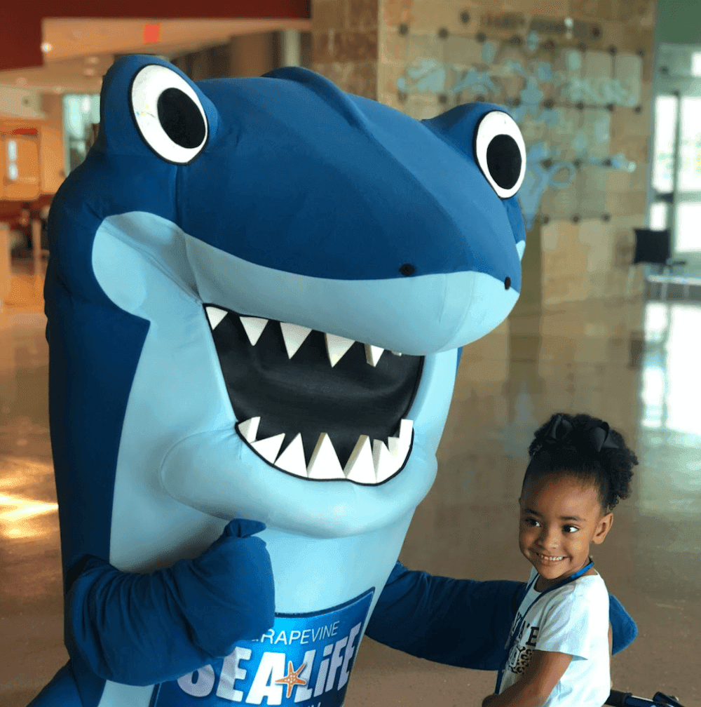 A SEA LIFE shark character kneels down to pose with a small child in a hospital who needs a frame to assist their walking.