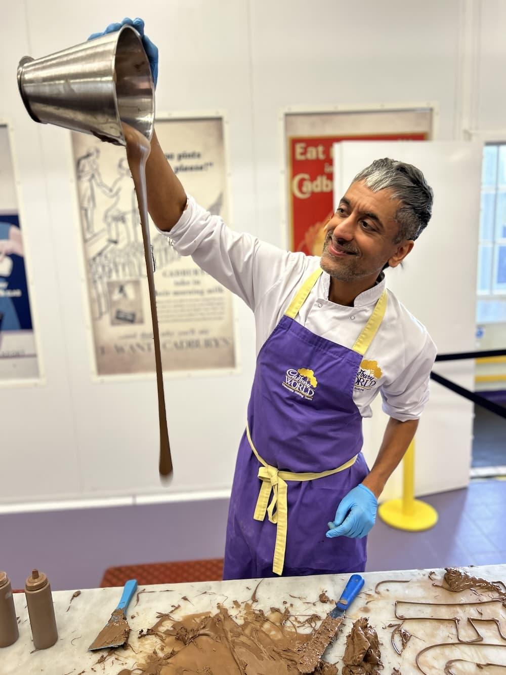 Man is wearing a white shirt and a purple apron with the text Cadbury World. He is holding a silver container in the air and is pouring melted chocolate onto a table.