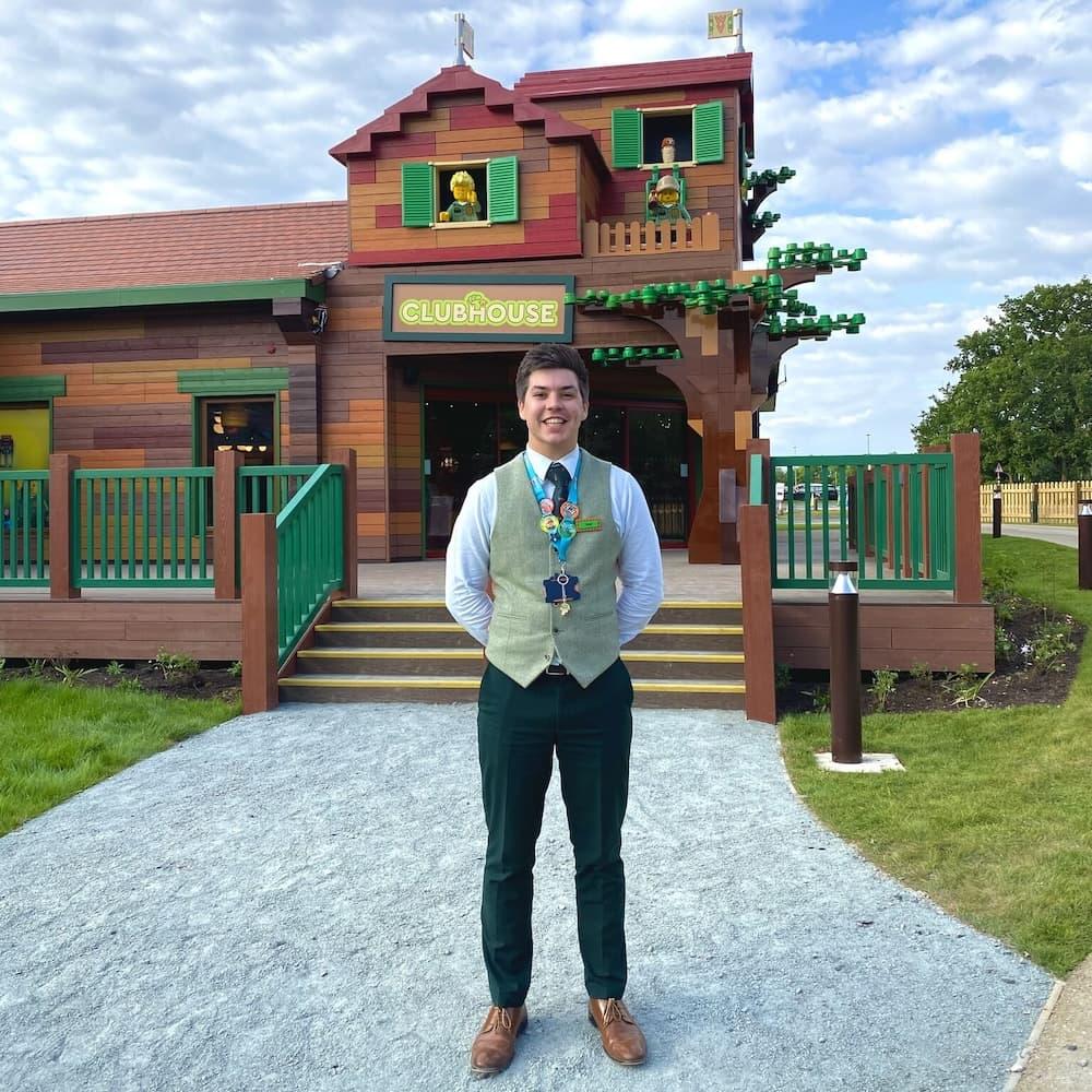 Man in brown brogue shoes, a white shirt and a grey waistcoat stands smiling in front of a LEGOLAND® brown LEGO® building.