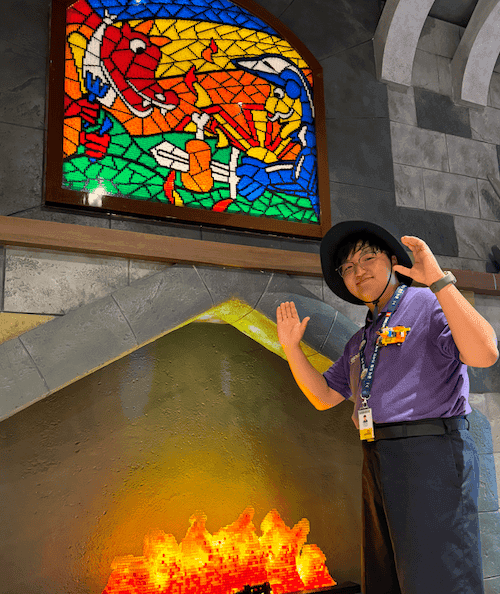 LEGOLAND® employee stands in front of a stone fireplace Above him is a stain glass window. He mimics a LEGO® man with his hands.