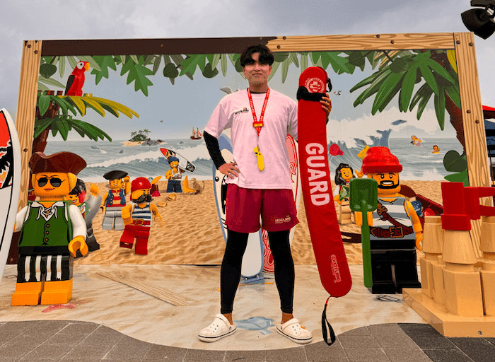 A lifeguard stands outside in front of a beach backdrop scene with LEGO® characters holding a red buoy that reads GUARD.