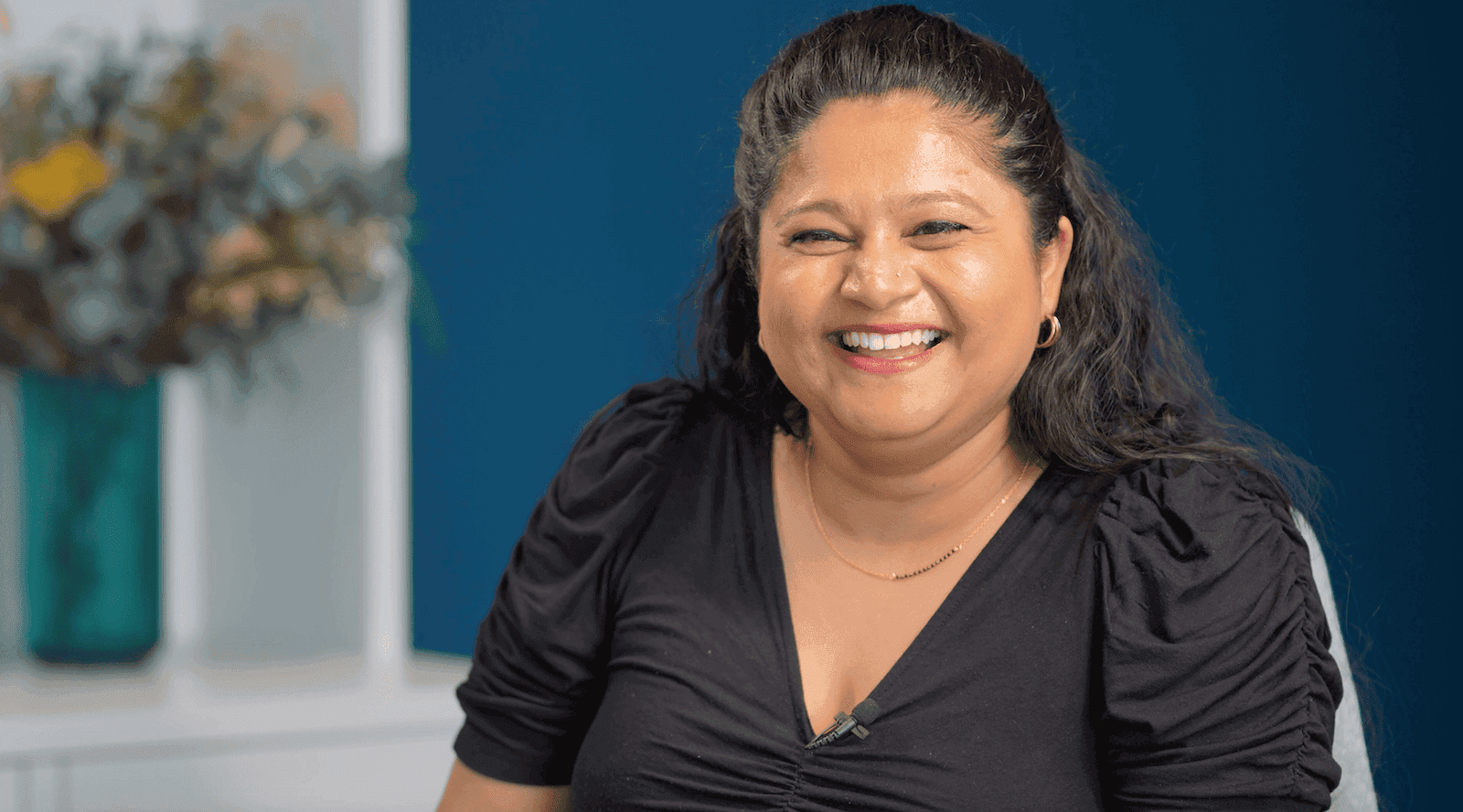 An image of a women laughing at the camera. She is sat on a chair and is wearing a black dress.