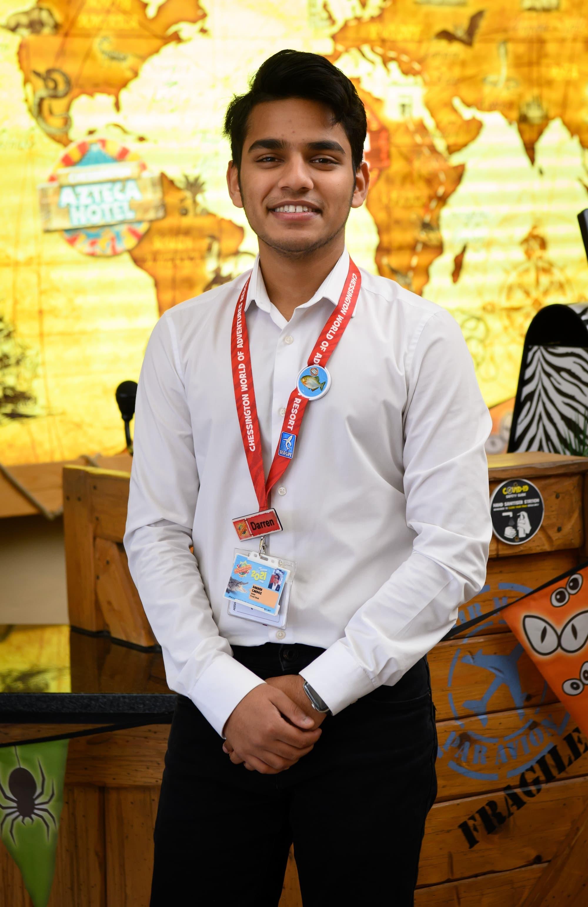 Man with brown hair wearing a white smart shirt with a red Chessington World of Adventures Resort lanyard over the top. There is a yellow and orange safari map on the wall in the background.