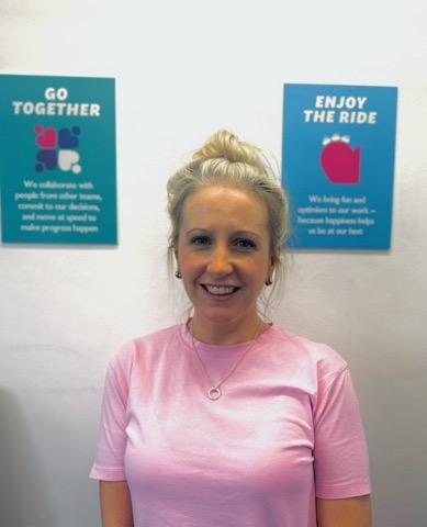 Blonde women has hair in a high bun, wearing a pink tshirt and silver necklace she is smiling at the camera. In the background there are two signs showing Merlin's values Go Together and Enjoy The Ride.