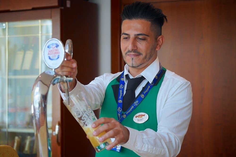 A man is working behind the bar at Gardaland. He is wearing a green waistcoat and is pouring a pint of beer.