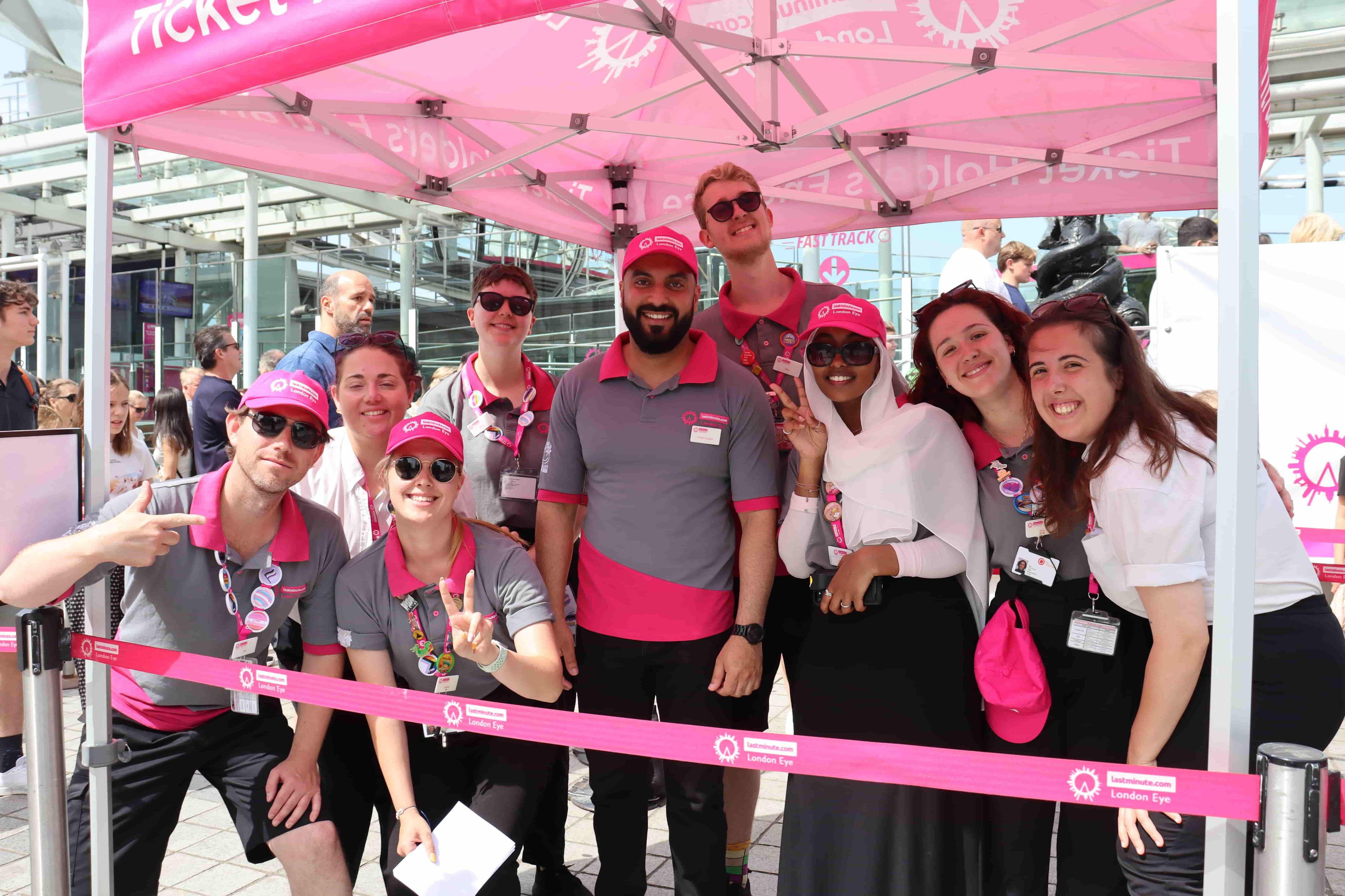 The London Eye team pose for a photo in the queue for the Eye. The group is a mixture of males and females in pink and grey uniforms.