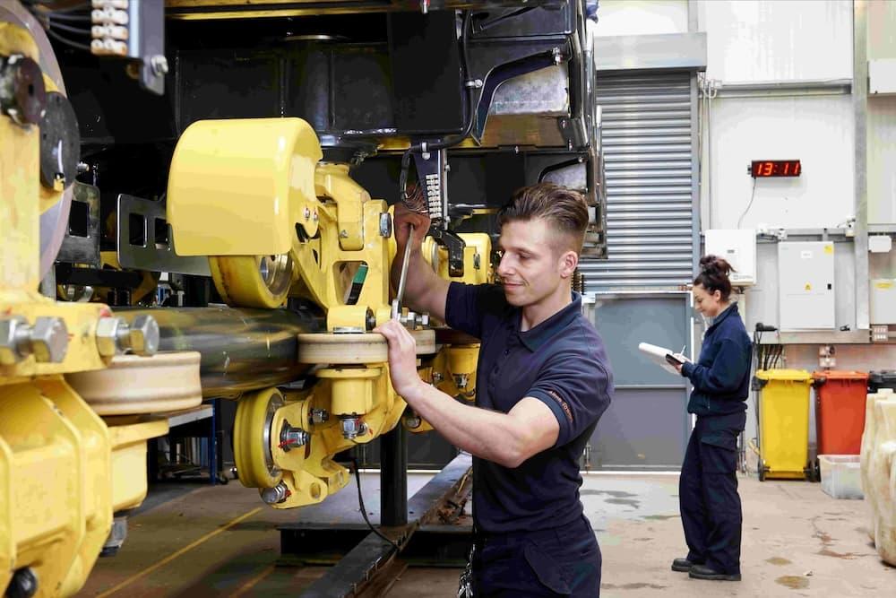 An engineer is inside a Merlin Entertainments factory fixing equipment for a rolleroaster. He is wearing a blue polo and is holding a spanner.