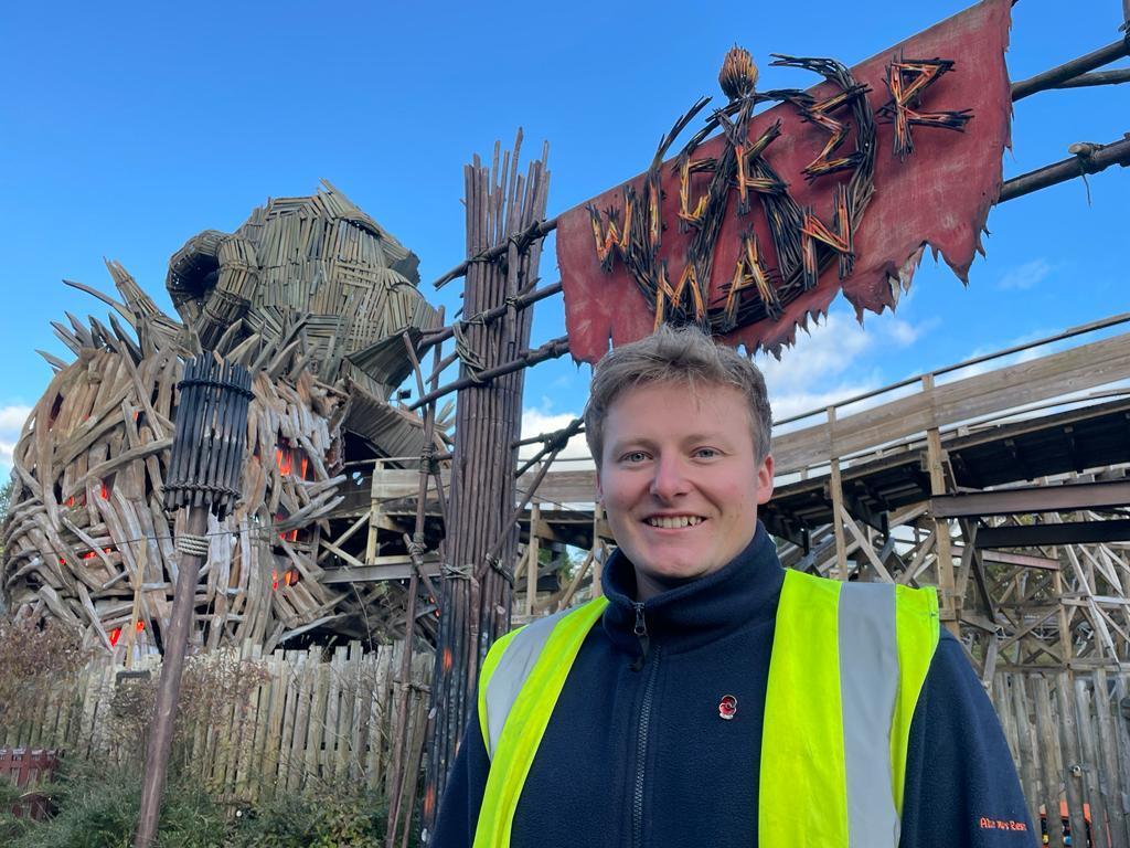 Man in a hi-vis jacket smiles in front of a sign that reads Wicker Man. Behind him and next to the sign is a large wooden ram.