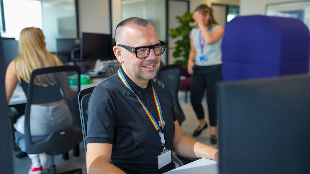 A man in glasses sits and smiles at his desk in the office looking at his computer. He is wearing a t-shirt and a rainbow lanyard.