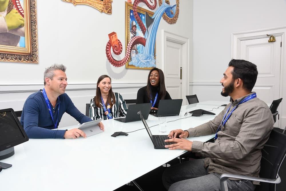 4 coworkers sit with laptops at a table smiling and looking at each other in a white room. On the wall is playful cartoon graphics.