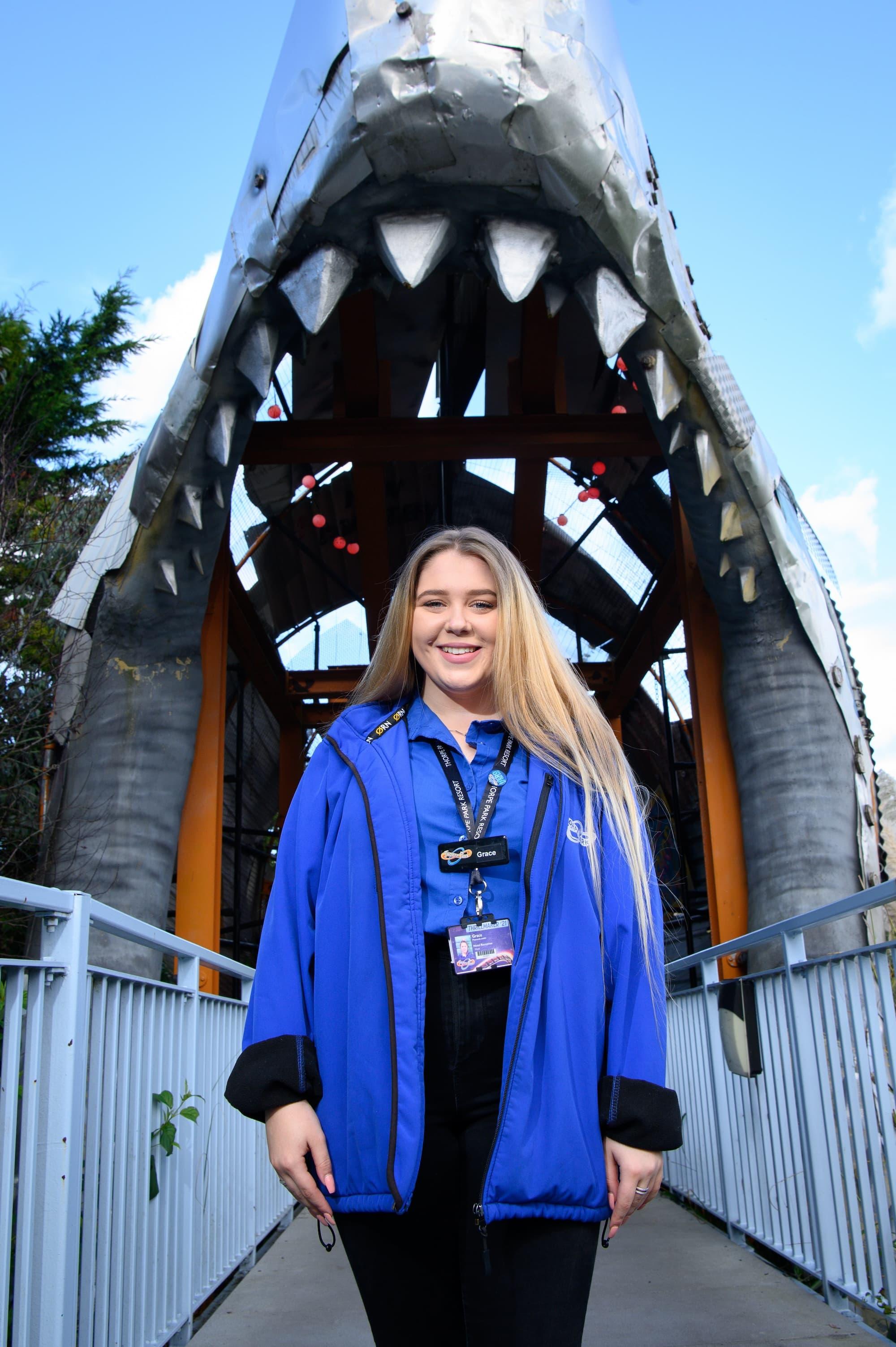 A blonde girl is stood on the bridge of Thorpe Park's entrance. She is wearing the staff uniform including a blue polo and a blue jacket with a lanyard.