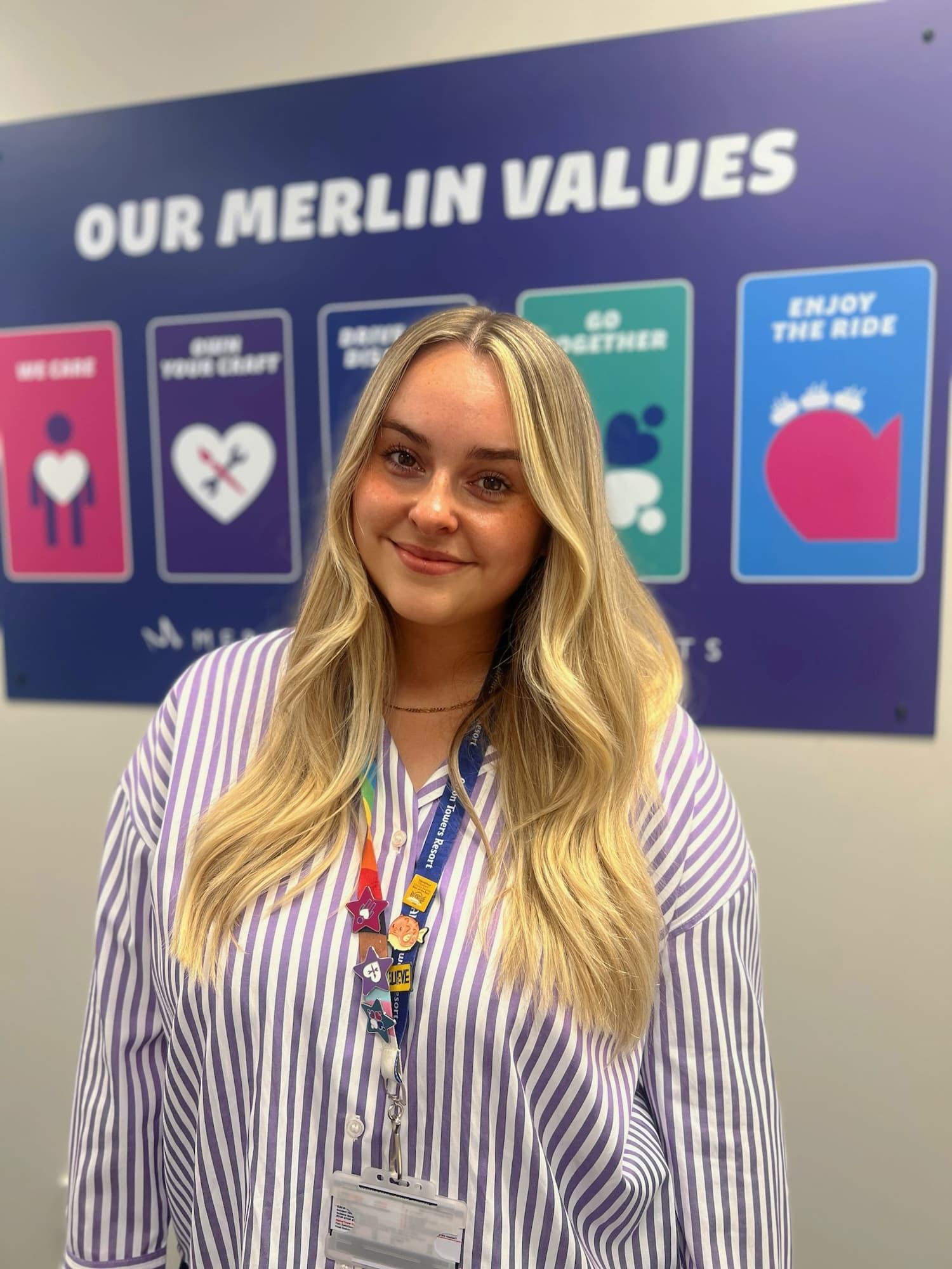 Women with long blonde hair is wearing a purple and white striped shirt smiling at the camera. Behind here there is a poster with the title 'Our Merlin Values' with cards for each of the values, she is also wearing a Merlin lanyard.