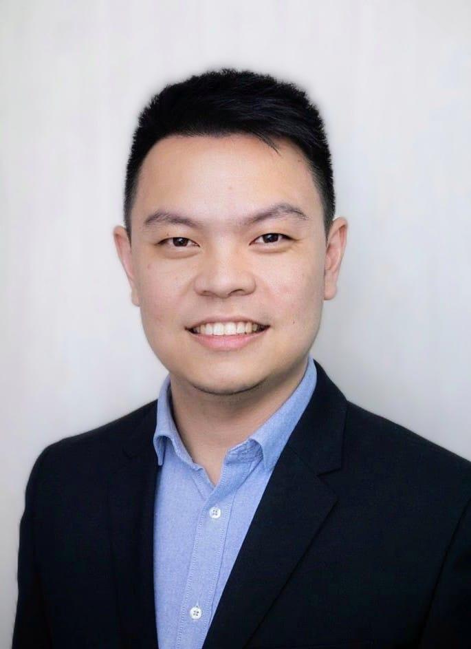 Man wearing a blue button up shirt and blazer is smiling for a headshot.