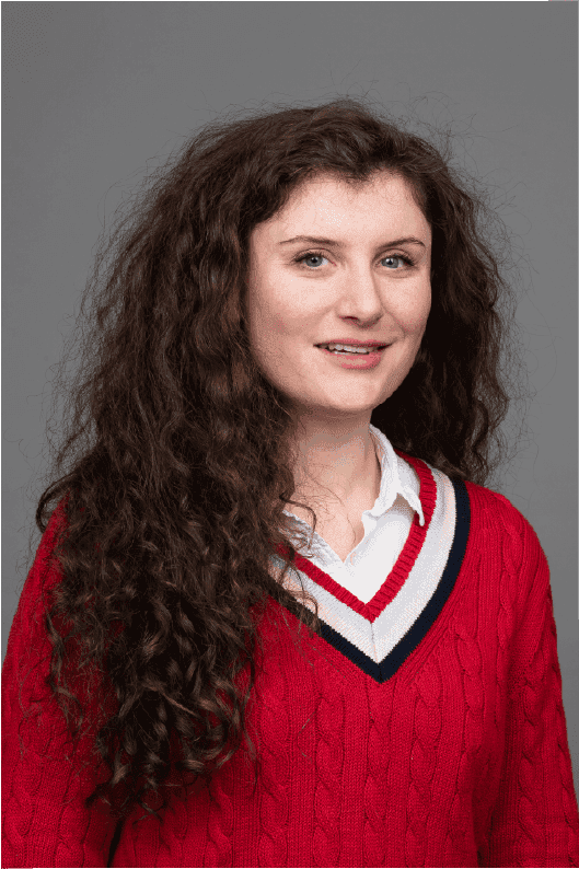 Women with long brown curly hair and a white polo shirt layered with red knitted sweatshirt is smiling.