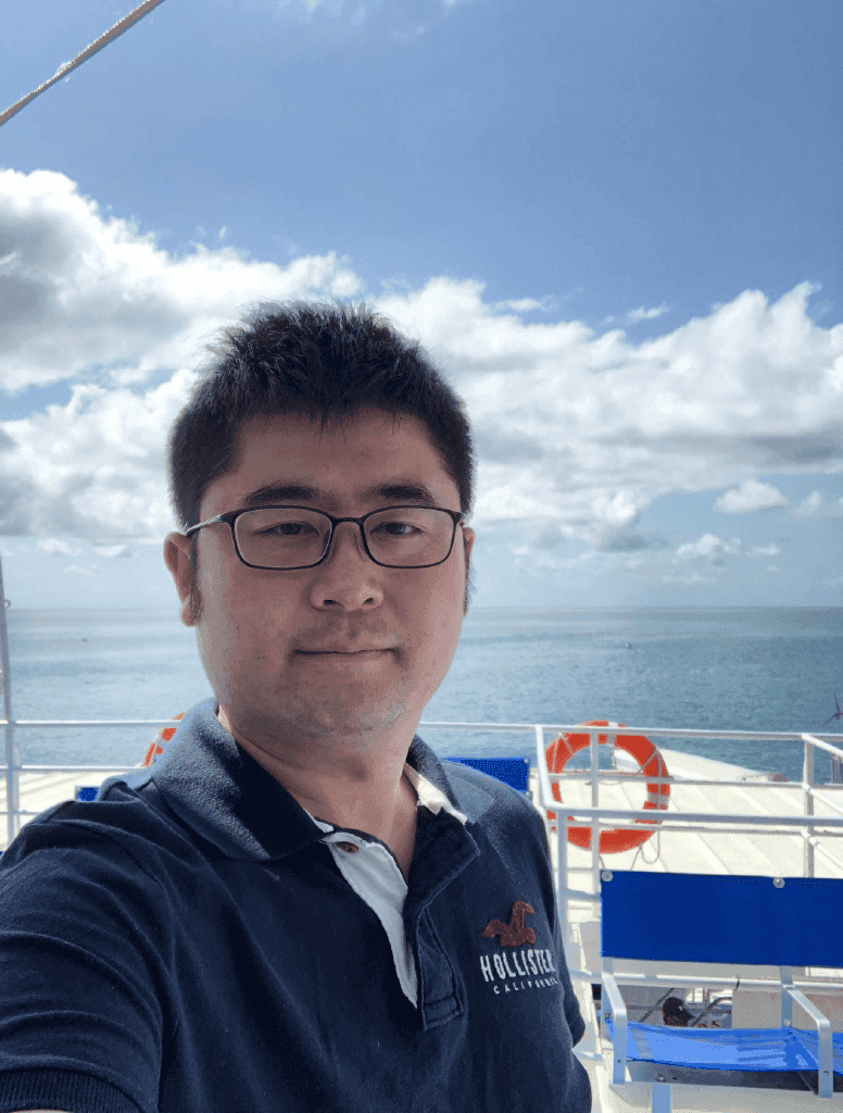 Man with short black hair is wearing a blue navy polo shirt and glasses stood on a boat with the sea line in the background.