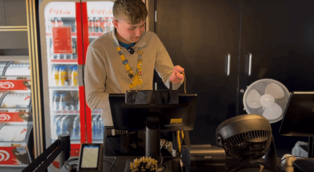 Man wearing a long sleeve beige polo top and yellow lanyard is packaging a gift into a black bag stood behind a counter.