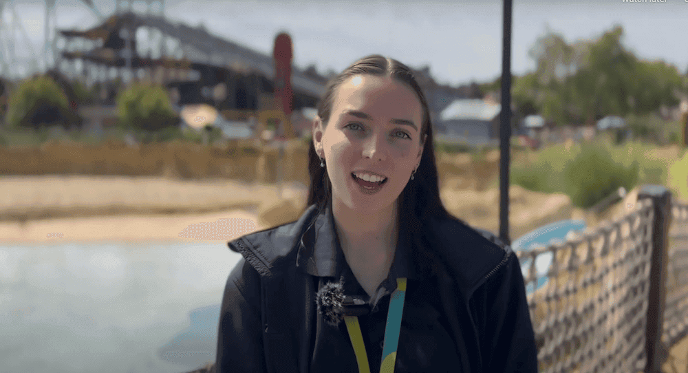 Women with brown long hair wearing a coat and Thorpe Park branded lanyard is stood on a bridge over a body of water. In the background there is a rollercoaster.