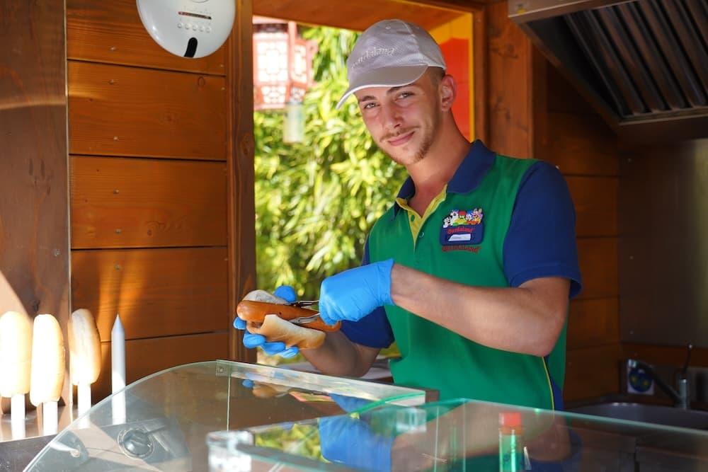 A man is wearing a green tshirt and a grey cap. He is wearing blue plastic gloves, holding tongs and putting a sausage into a bun.