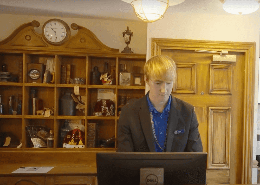 Young man standing behind a hotel reception desk typing on a computer.