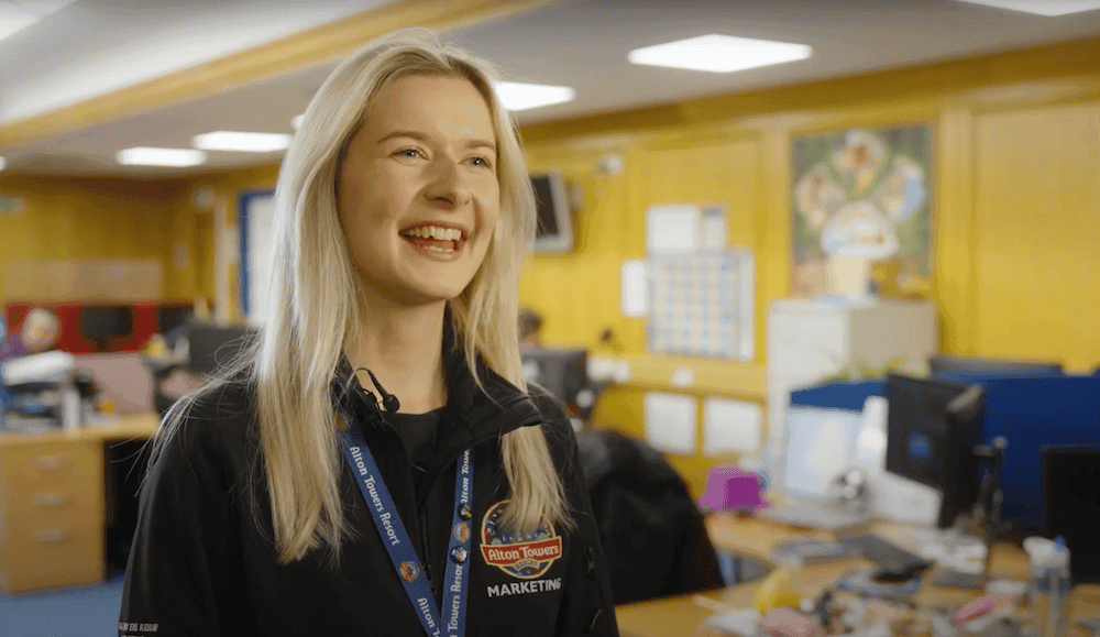 Close up shot of a woman in a black outfit with Alton Towers Resort branding, smiling in a yellow office.