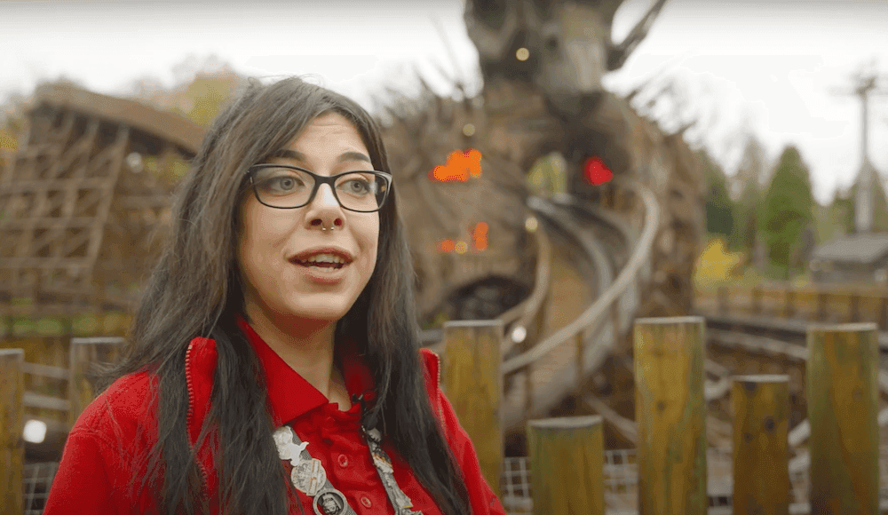 Woman in a red outfit standing in front of a wooden rollercoaster with fire effects.