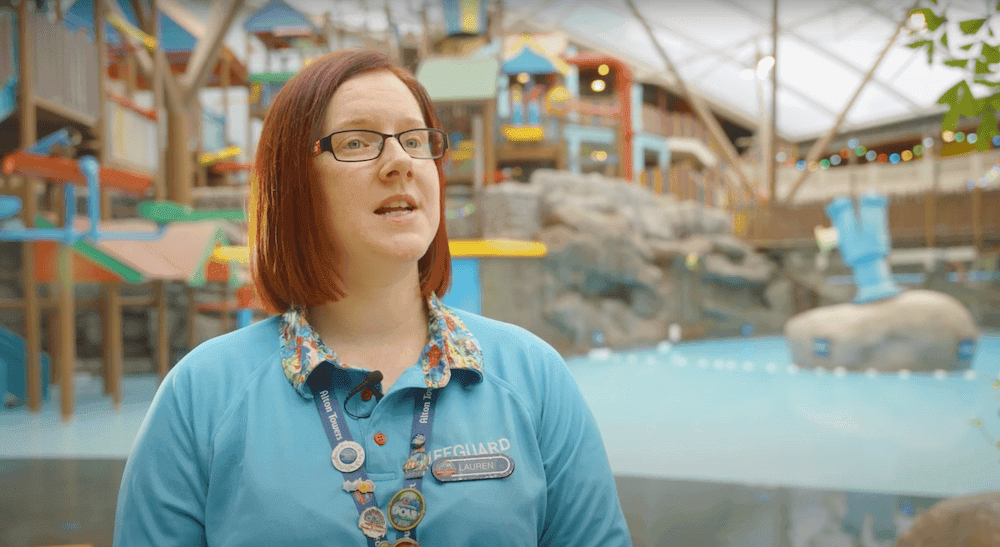 Woman in a blue polo shirt and lanyard standing in front of an indoor waterpark with swimming pool and obstacles.