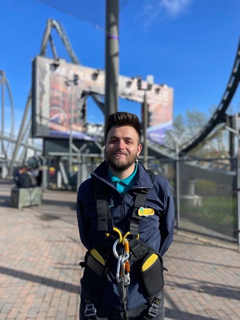 Man wearing a blue coat and skyline equipment stood infront of a rollercoaster.