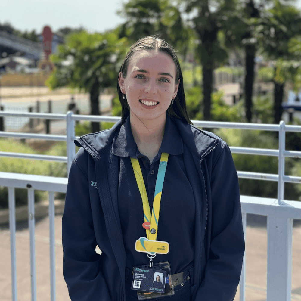 Women with brown hair wearing blue jacket and yellow Thorpe Park lanyard stood in front of a white metal fence.