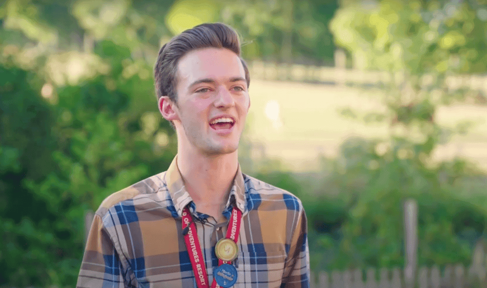 Man in a blue and brown checked shirt, standing infront of a tree filled field.