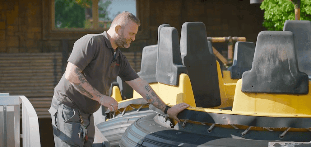 A male mechanic in a grey outfit is leaning on a yellow inflatable water-ride boat