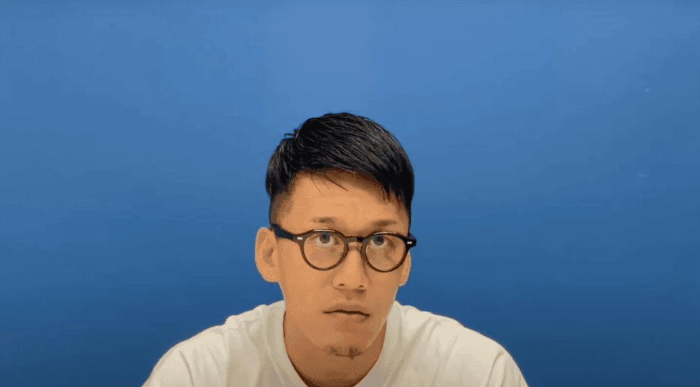 Man wearing black circular frame glasses in a white t-shirt sat infront of a blue background.