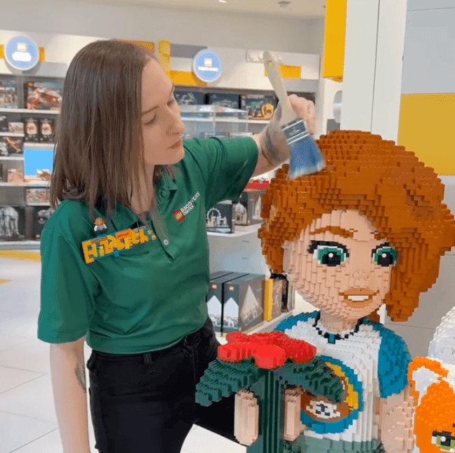 Women in a green polo shirt is holding a brush and brushing the auburn hair of a LEGO® model.