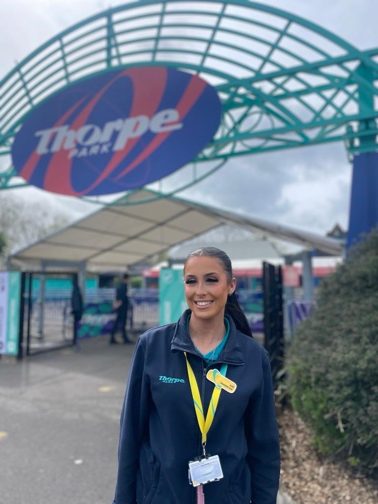 Women in blue jacket and yellow lanyard and hair in a slicked back ponytail is stood in front of a Thorpe Park sign.