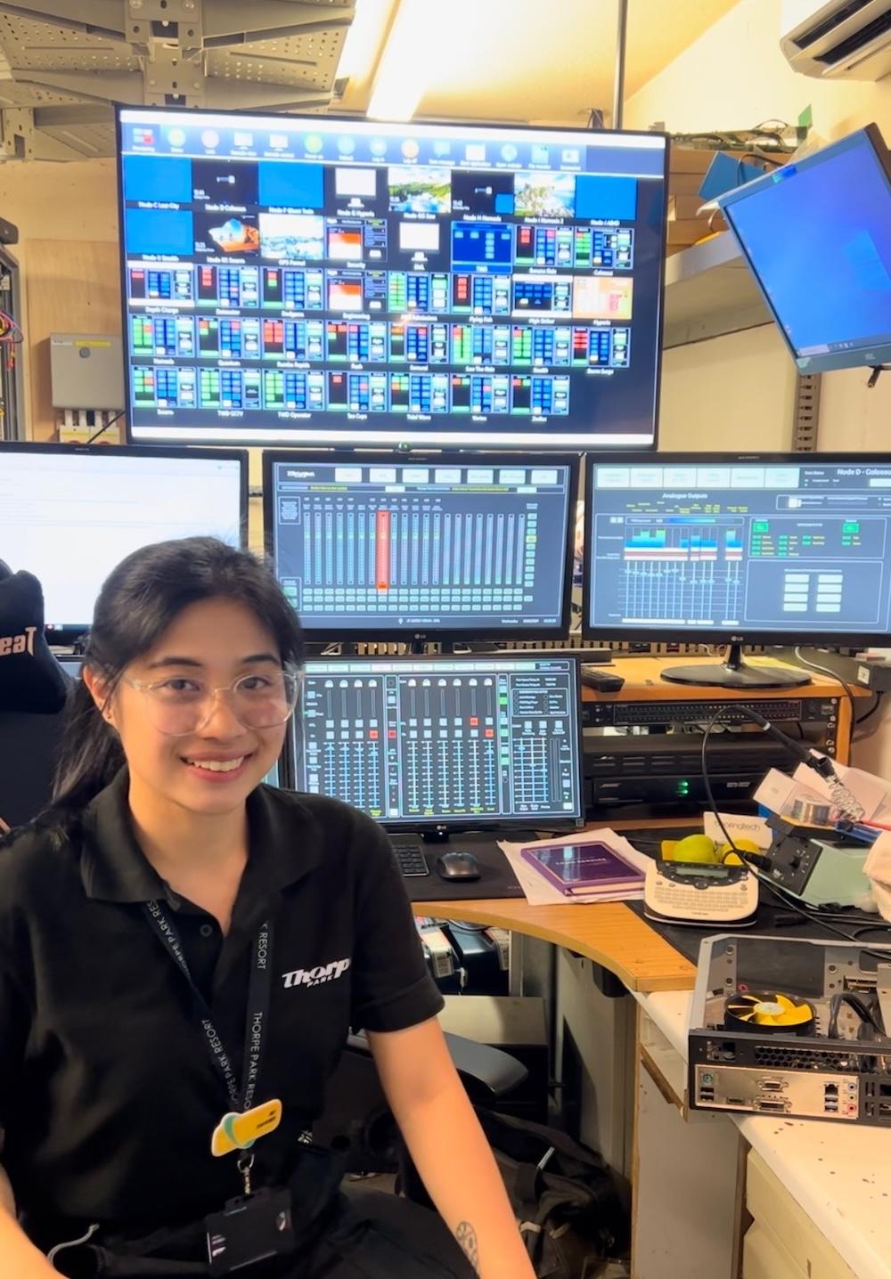 Women with long hair in a ponytail and wearing a blue polo shirt is sat on a chair with a selection of monitor screens behind her.