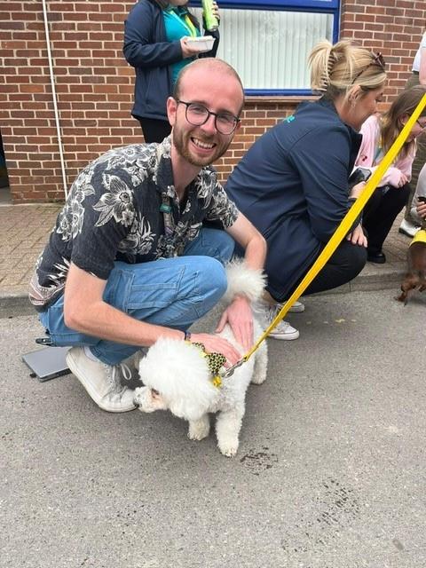 Man wearing a patterned shirt is crouched on the floor stroking a white dog that is stood on the pavement.